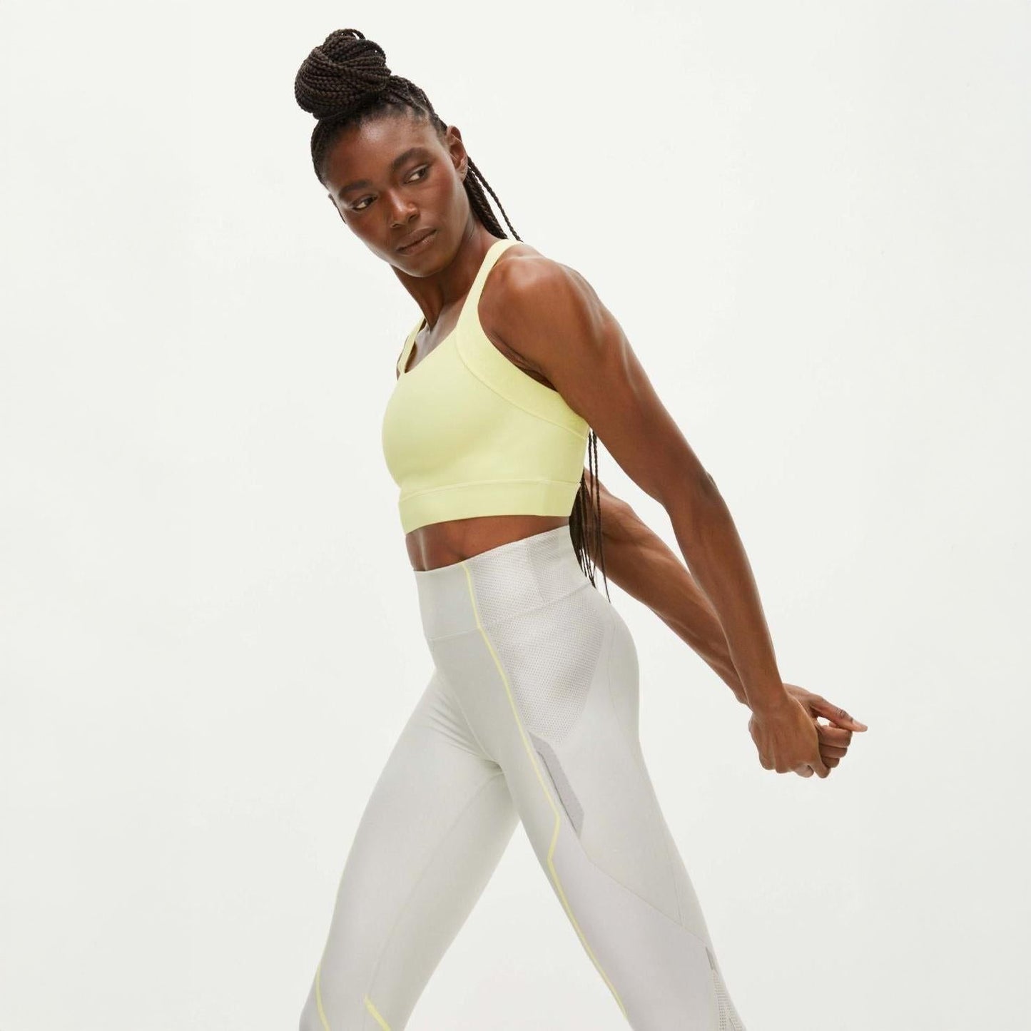 A person in a yellow Rohnisch Kay Sports Bra made from recycled polyester and light gray leggings stretches their arms behind their back against a plain background.