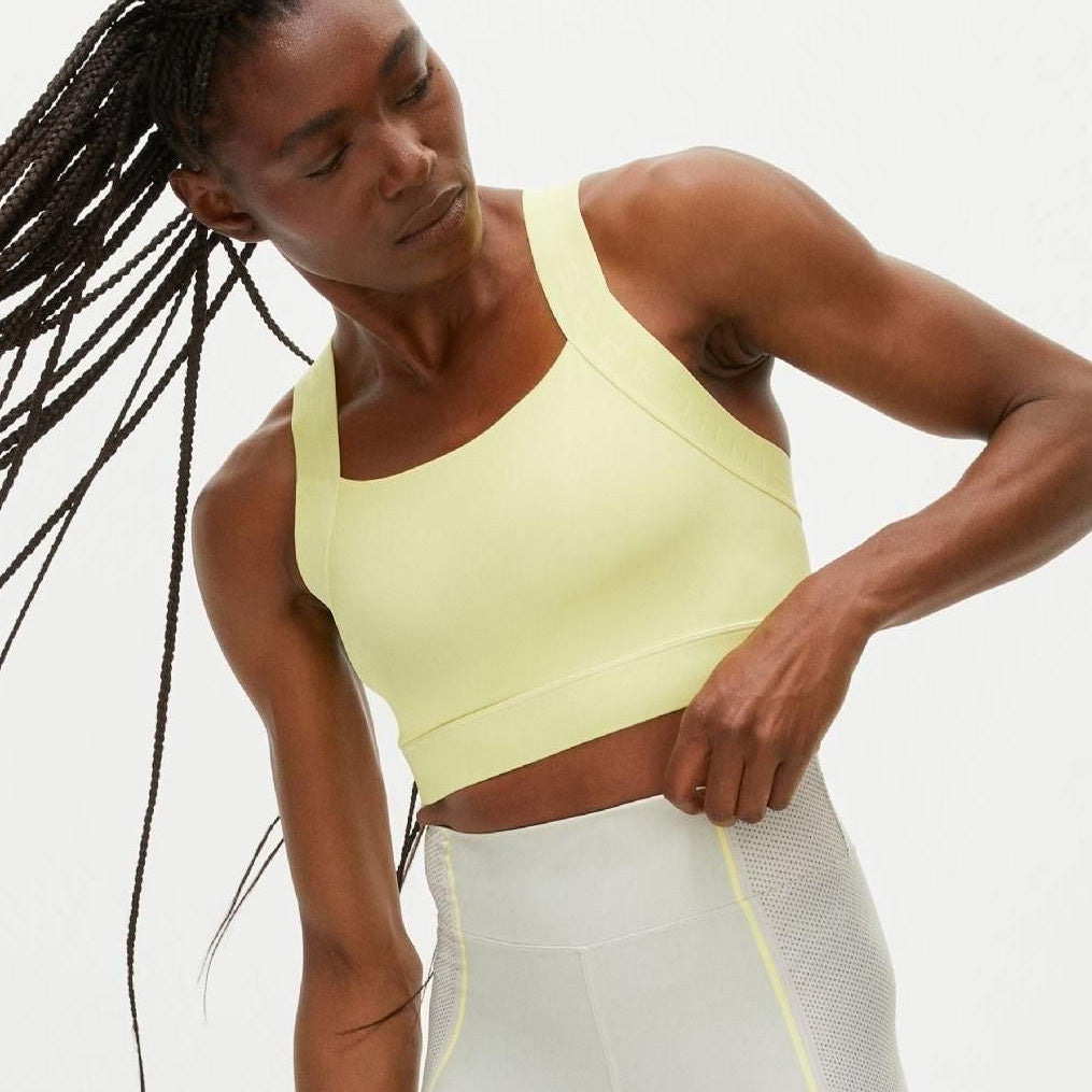 A person with long braids wears a Rohnisch Kay Sports Bra and leggings, crafted from moisture-wicking recycled polyester, standing against a plain background.