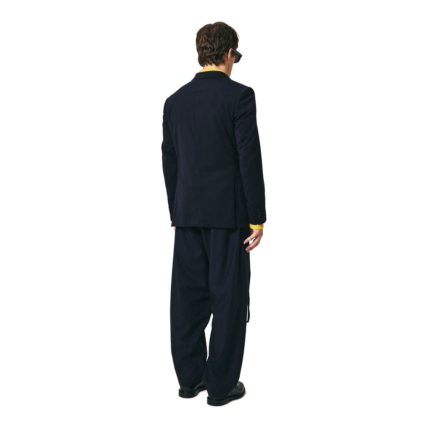 A man wearing a dark suit and yellow shirt, possibly a trendy Seven Gauge Double Breast Blazer Tropical Wool, Blu, stands with his back turned, facing away from the camera. He is also wearing dark sunglasses.