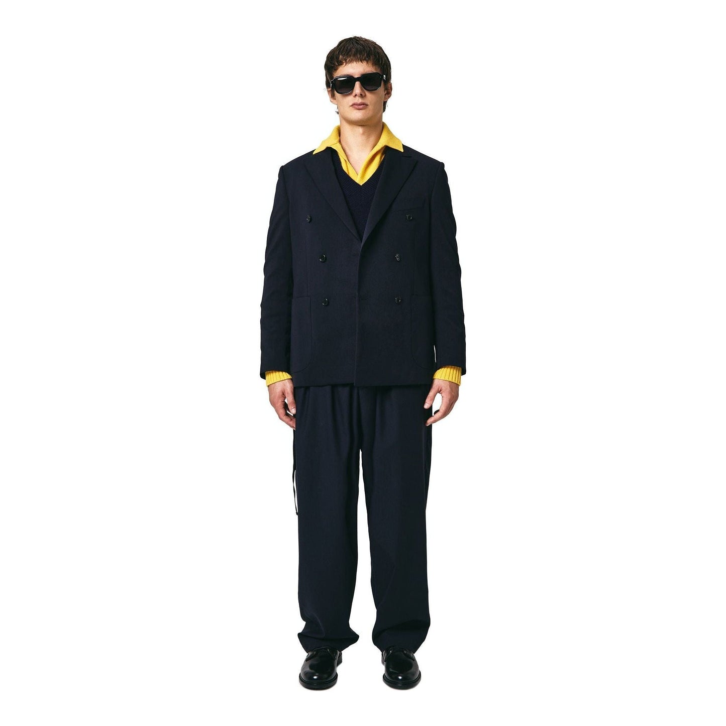 A person stands against a white background wearing a Double Breast Blazer Tropical Wool, Blu by Seven Gauge over a yellow shirt, loose-fitting dark pants, shiny black shoes, and sunglasses.