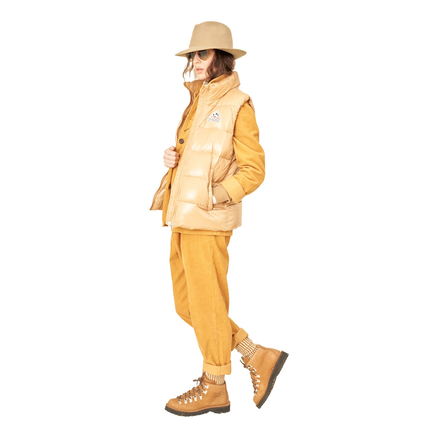 A person in a tan outfit showcases Universal Works Military Chino Corduroy trousers, a puffer vest, boots, sunglasses, and a wide-brimmed hat while standing in profile.